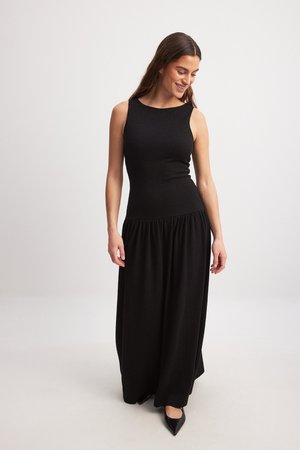 Black maxi dress with sleeveless design, textured fabric, fitted bodice, and gathered waistline. Worn with black pointed heels.