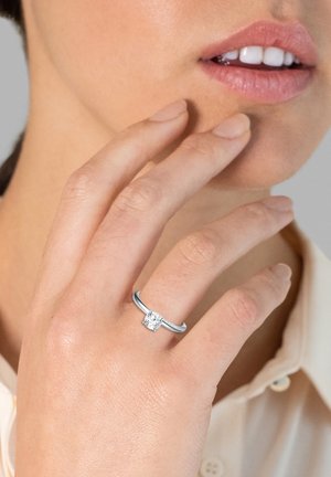 Silberner Ring mit einem quadratischen Diamantmittelstein, der eine glatte Ringschiene und kleinere Akzentdiamanten entlang des Schenkels aufweist, auf einer Hand präsentiert.