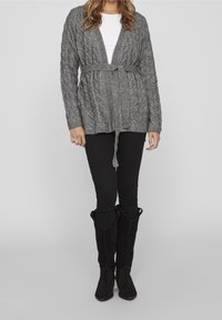 Šedý pletený kardigan s uvázaným pasem, spárovaný s černými skinny džíny a černými semišovými botami po kolena. Hladká textura, otevřený střih.