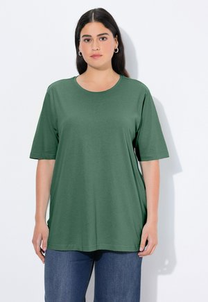 Donna con capelli scuri che indossa orecchini a cerchio d'argento, una maglietta verde ampia a maniche corte e jeans blu, in piedi davanti a uno sfondo bianco.