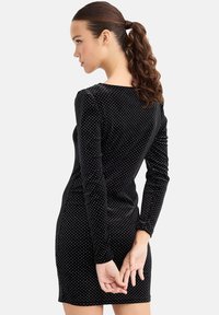 Vestido de terciopelo negro con mangas largas, con lunares blancos, escote redondo y silueta ajustada, que muestra una textura suave.