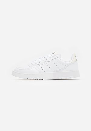 Biały niski sneaker z perforowanymi detalami po bokach, białymi sznurowadłami i grubą gumową podeszwą, przedstawiony w profilu z prawej strony na jednolitym tle.