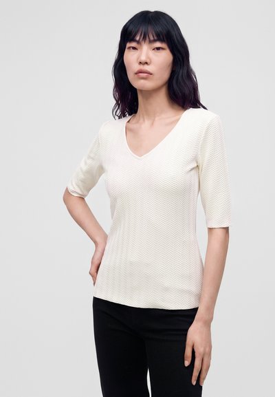 Femme aux cheveux noirs mi-longs portant un haut blanc texturé à manches courtes et un pantalon noir, debout devant un fond gris uni.