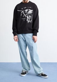 Sudadera negra con gráfico blanco de un oso y un perro, texto "SÉ AGRADABLE CONMIGO, POR FAVOR," combinada con jeans holgados de color azul claro y zapatillas negras.