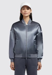 Jachetă bomber gri din satin cu manșete ribbate. Dispune de un fermoar frontal, buzunare laterale și o textură netedă și lucioasă. Purtată cu pantaloni de trening gri asortați.