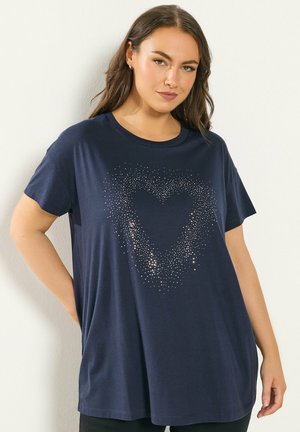 Kvinde iført navyblå T-shirt med glitrende hjerte-design foran, stående mod hvid baggrund med hånden på hoften.