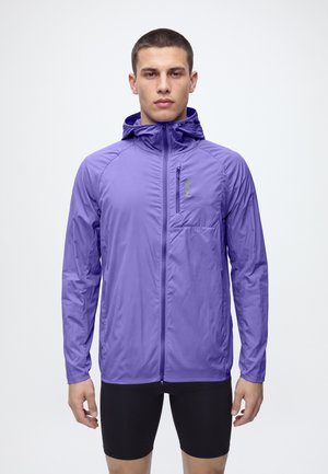Un homme porte une veste violette à capuche et à fermeture éclair, avec une poche poitrine, ainsi qu’un cuissard de cyclisme noir, sur un fond blanc uni.