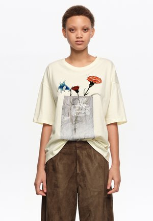 Persona vistiendo una camiseta crema oversize con estampado floral y un gráfico de bolsa de papel, combinada con pantalones marrones sueltos, de pie contra un fondo blanco.