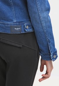 Džínová bunda v tmavě modré barvě s knoflíkovým zapínáním a štítkem na zadní straně. Černé kalhoty se hladkým povrchem a diskrétní kapsou.