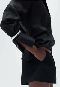 Schwarzes Seidenshirt mit hochgekrempelten Ärmeln, das mit einem runden silbernen Akzent versehen ist. Kombiniert mit hochgeschnittenen schwarzen Shorts, die klare Linien und Texturen betonen.