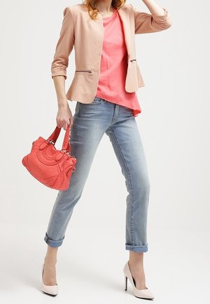 Koraalkleurige handtas met textuurdetails, gecombineerd met een lichtroze blazer, een roze shirt en lichtblauwe jeans met opgerolde mouwen. Witte hoge hakken.