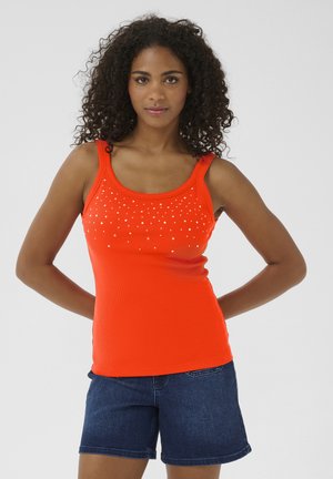 Femme aux cheveux bouclés portant un débardeur orange à petits points blancs et un short en jean bleu foncé, debout les mains derrière le dos.