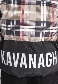 Nafukovací bunda s béžovým, čiernym a červeným károvaným vzorom, s čiernou spodnou časťou, na ktorej sa nachádza veľký biely text "KAVANAGH" na leme.