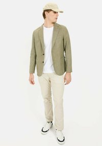 Licht olijfblazer met twee knopen, gecombineerd met een wit T-shirt en beige broek. Gekenmerkt door een beige pet en witte sneakers met zwarte accenten.