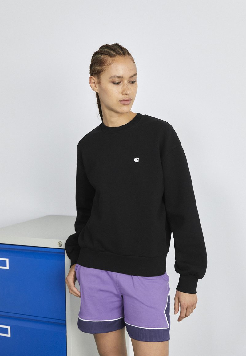 Schwarzer Sweatshirt mit rundem Hals, langen Ärmeln und einem kleinen weißen Logo auf der Brust, kombiniert mit lila Shorts mit weißem Abschluss.