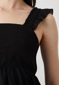 Rochie neagră cu un corsaj texturat și detalii fronsate, având bretele cu volane. Țesătura este ușoară și fluidă.