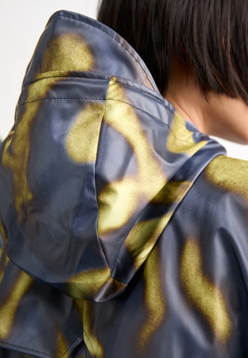 Veste à capuche avec une texture lisse et brillante. Présente un motif multicolore de bleus foncés et de nuances dorées jaunâtres. Coutures visibles en accent.