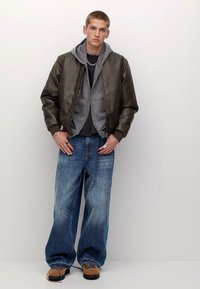 Overmaatspantalons van denim, een grijze hoodie onder een donkergroene bomberjack, met ribbelmanchetten en taupe schoenen met zwarte zolen.