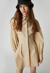 Beige Hemdkleid mit tailliertem Oberteil, plissiertem Rock und langen Puffärmeln. Verfügt über einen klassischen Kragen und eine durchgehende Knopfleiste.