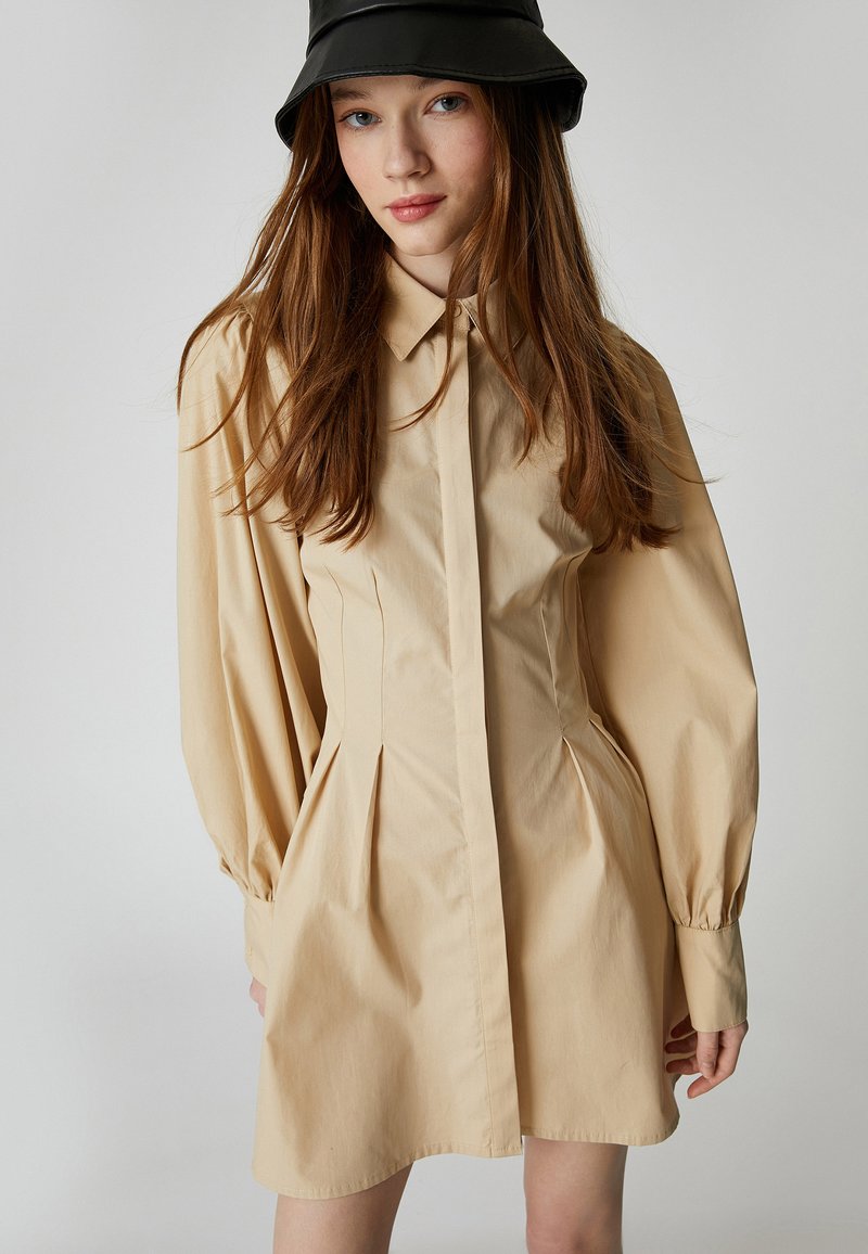 Beige Hemdkleid mit tailliertem Oberteil, plissiertem Rock und langen Puffärmeln. Verfügt über einen klassischen Kragen und eine durchgehende Knopfleiste.