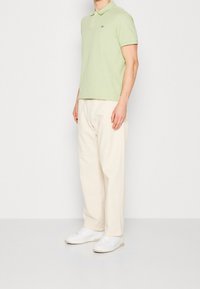 Hellgrünes Polohemd mit kurzen Ärmeln und gesticktem Logo. Locker sitzende cremefarbene Hose, weiße Sneaker, minimalistisches Design.