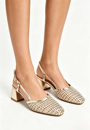 Femme portant des chaussures à talons blocs tissées dorées à bride arrière sur fond blanc, montrant les vues avant et latérale des chaussures.