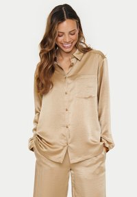 Beige satin button-up shirt with a chest pocket, long sleeves, and a relaxed fit. Features a smooth texture and slightly rounded hem.