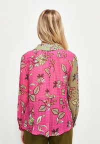 Blusa floreale con uno sfondo rosa vibrante e accenti verdi. Maniche lunghe, colletto e intricate trame di foglie e fiori.