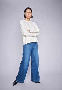 Witte sweatshirt met een relaxte fit, gecombineerd met brede blauwe denim jeans. Model draagt zwarte puntige hakken. Eenvoudig, strak ontwerp.