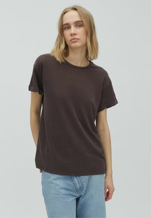 Jonge vrouw met schouderlang blond haar, gekleed in een donkerbruin T-shirt met korte mouwen en lichtblauwe spijkerbroek, staand tegen een effen grijze achtergrond.