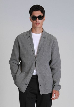 Grijze geribbelde gebreide cardigan met geknoopte revers, een enkele borstzak en twee voorzakken. G gedragen over een witte t-shirt en zwarte broek.