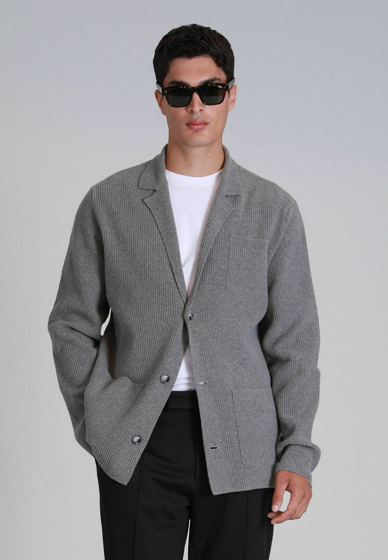 Grijze geribbelde gebreide cardigan met geknoopte revers, een enkele borstzak en twee voorzakken. G gedragen over een witte t-shirt en zwarte broek.