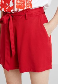 Pantalones cortos rojos de tejido suave, con un cinturón atado en la cintura, bolsillos laterales y un corte relajado con detalles de dobladillo cosido.
