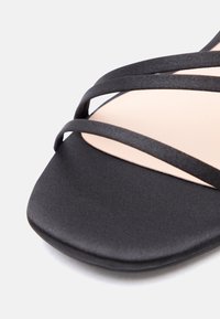 Sandale à bout carré noir avec fines lanières en tissu croisées et semelle beige, vue en gros plan sur fond blanc.