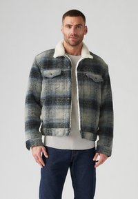 Chaqueta de mezclilla a cuadros con una paleta de colores azul, gris y crema, que presenta un cuello de borreguillo, dos bolsillos en el pecho y un corte corto.