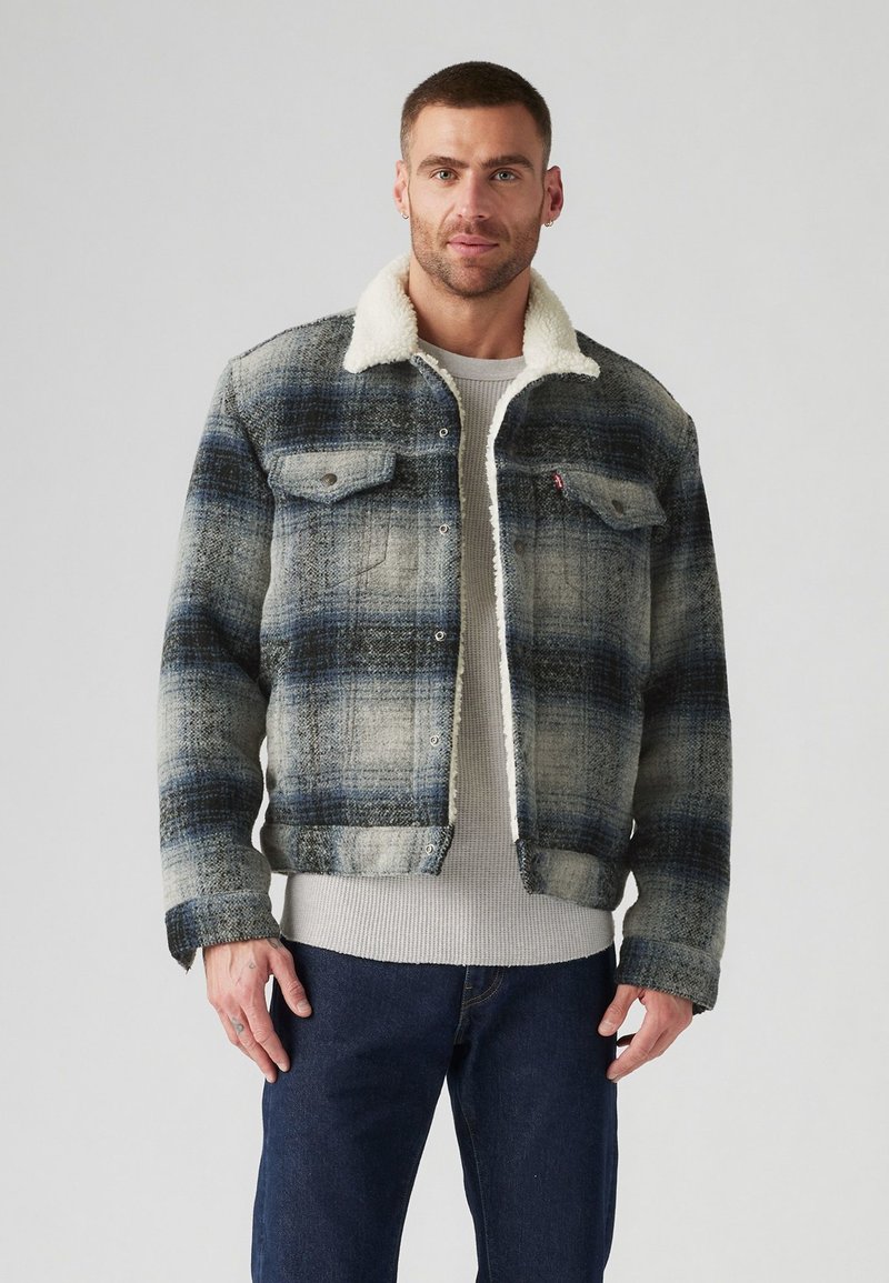 Chaqueta de mezclilla a cuadros con una paleta de colores azul, gris y crema, que presenta un cuello de borreguillo, dos bolsillos en el pecho y un corte corto.