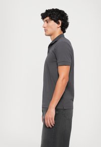 Young man with curly hair stands side-facing, wearing a dark gray short-sleeve polo shirt and faded black jeans against a plain background.