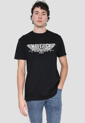 Young man with curly dark hair wearing a black "Maverick" graphic t-shirt and blue jeans, standing against a plain light gray background.