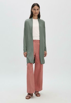 Lange, open voorkant cardigan van groene stof, gecombineerd met lichtroze wijde broek. Eenvoudig wit shirt eronder, platte bruine sandalen.