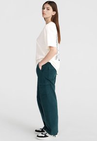 Off-white oversized t-shirt met een grafische print op de achterkant, gecombineerd met teal cargo pants met meerdere zakken en een textuurstof.