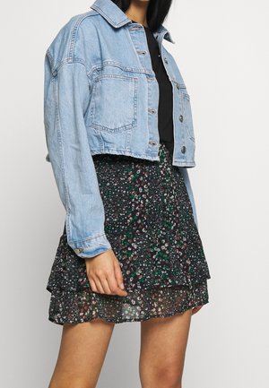 Femme portant une veste en denim courte bleu clair sur un haut noir avec une jupe florale noire à plusieurs couches ornée de fleurs vertes et blanches.