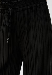 Zwarte geplooide broek met een trekkoord in de taille, gemaakt van een textuurstof en met metalen accenten op de koorden. Close-up van het materiaal.