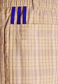 Harmaa ruudullinen shortit, joissa on violettia korostusraitaa, elastinen vyötärö ja näkyvä kirjailtu brändilogo taskussa.