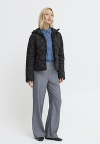 Femme blonde portant une veste matelassée noire par-dessus une chemise en denim bleu, avec un pantalon large gris et des chaussures noires, se tenant sur fond blanc.