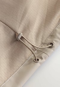 Tissu beige avec une texture en maille, comportant un cordon de serrage avec un arrêt en plastique et des œillets assortis.