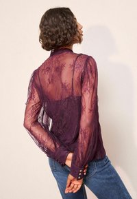 Žena s krátkymi kučerami, oblečená v priesvitnej purpurovej čipkovanej blúzke a modrých džínsoch, stojaca s rukami zovretými za chrbtom.