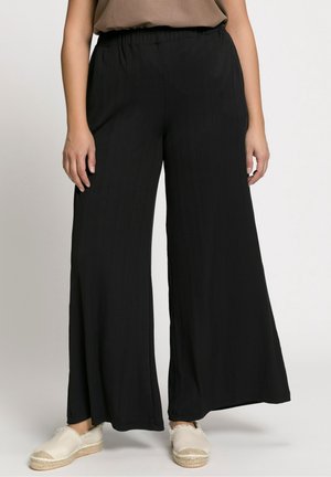 Kobieta ubrana w luźne czarne spodnie o szerokich nogawkach z elastycznym pasem i beżowe espadryle na jasnym tle.