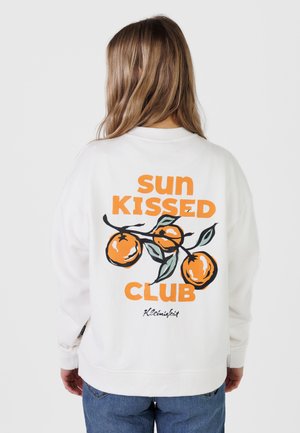Person mit langen Haaren von hinten gesehen, trägt ein weißes Sweatshirt mit orangefarbenem Text "sun kissed club" und illustrierten Orangen auf dem Rücken.