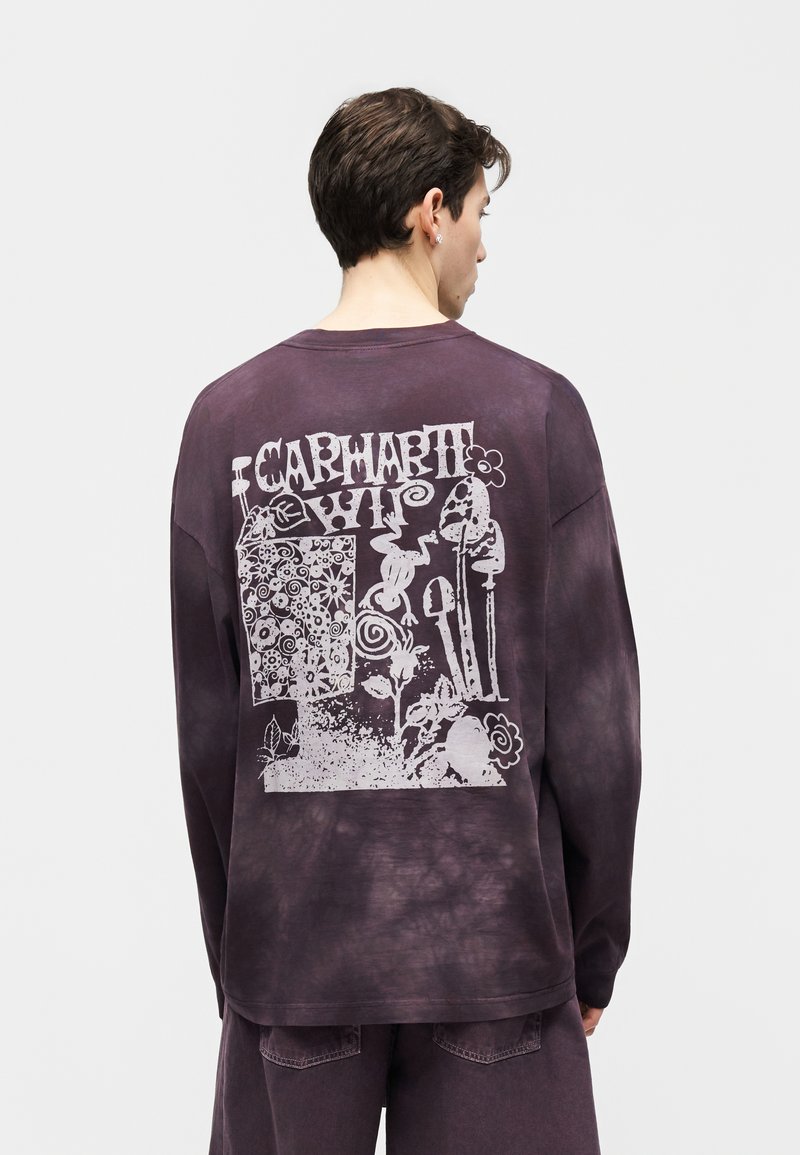 Uomo che indossa una maglia a maniche lunghe tie-dye viola scuro con grafica floreale astratta bianca e scritte sul retro, in piedi di spalle.