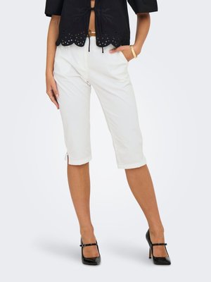 Femme portant un pantalon blanc jusqu'aux genoux, un top court noir et des chaussures noires à talons hauts, debout avec une main dans la poche.